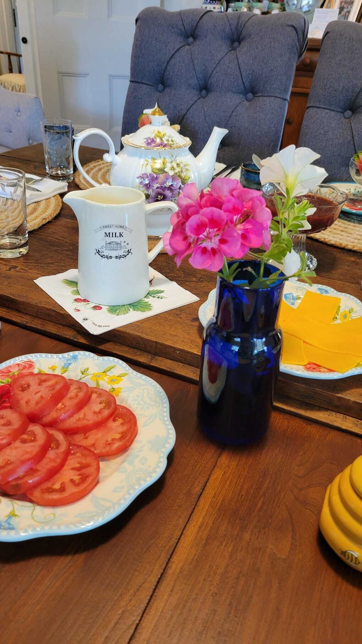 Une table à manger avec une assiette de tomates coupées en tranches, un pichet étiqueté « LAIT DE MAISON », un vase bleu rempli de fleurs et une théière décorée de motifs floraux.