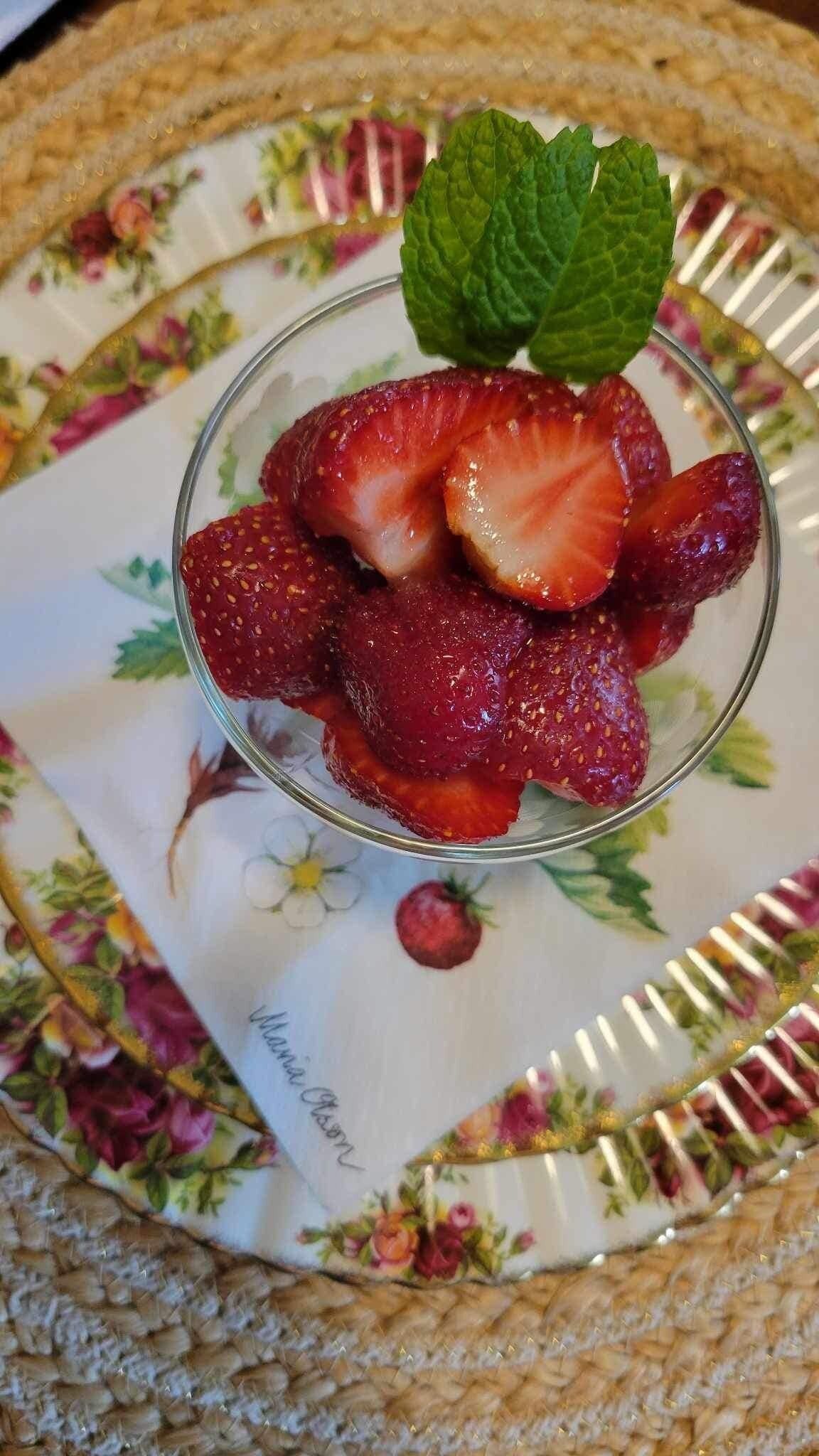 A glass bowl with sliced strawberries and a mint leaf on a floral-patterned plate.