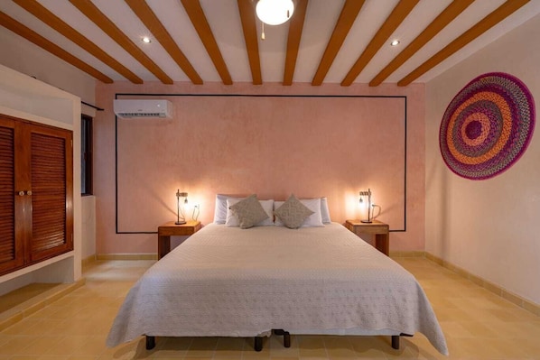 A bedroom with a large bed, wooden ceiling beams, two bedside lamps, a wall-mounted air conditioner, and a colorful circular wall decoration. - Casa Pakal (Valladolid)