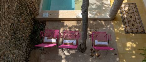 Un área junto a la piscina con tres sillas reclinables rosadas, un árbol y un suelo de baldosas con diseños decorativos.