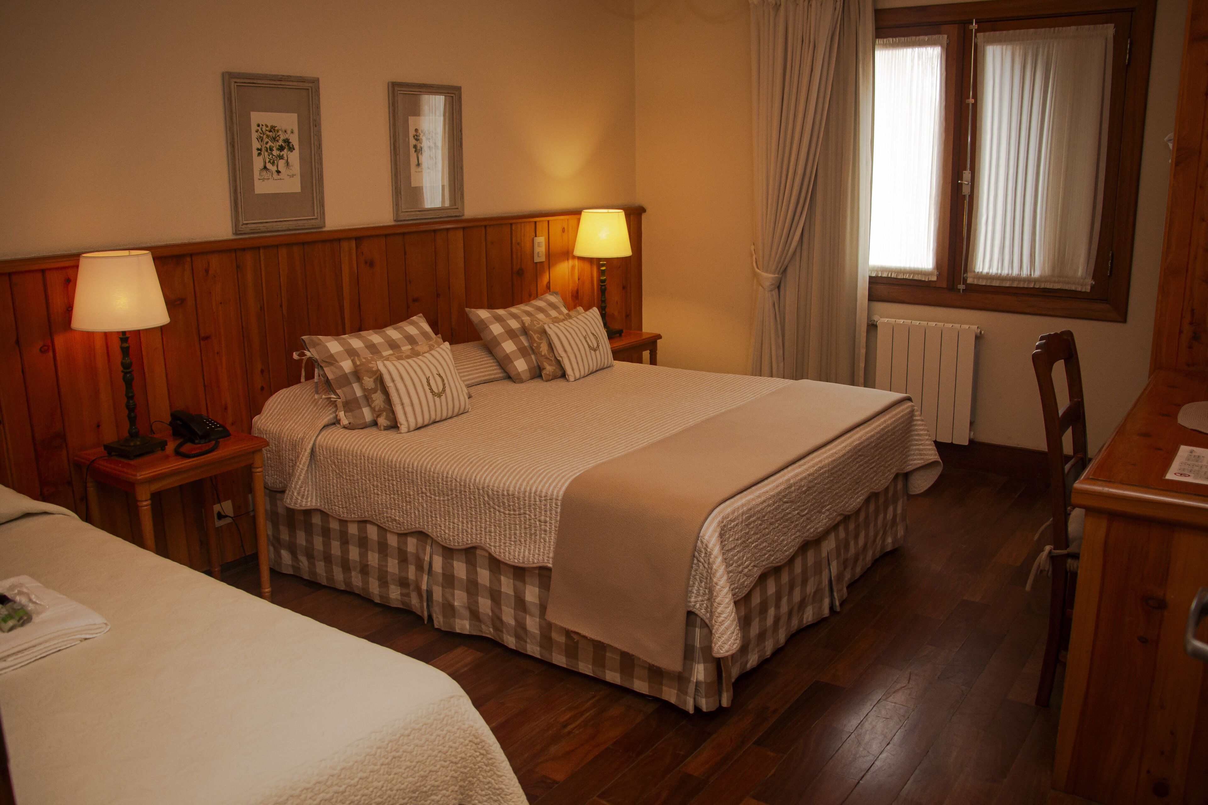 A hotel room with a large bed, two bedside tables with lamps, a wooden headboard, a window with curtains, and a radiator.