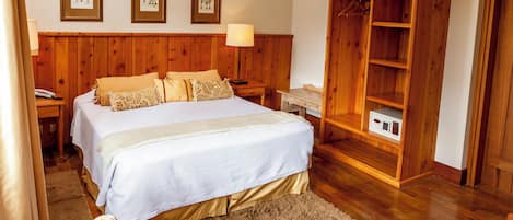 Un dormitorio con cabecera de madera, cama con ropa de cama color beige, mesita de noche con lámpara y armario de madera.