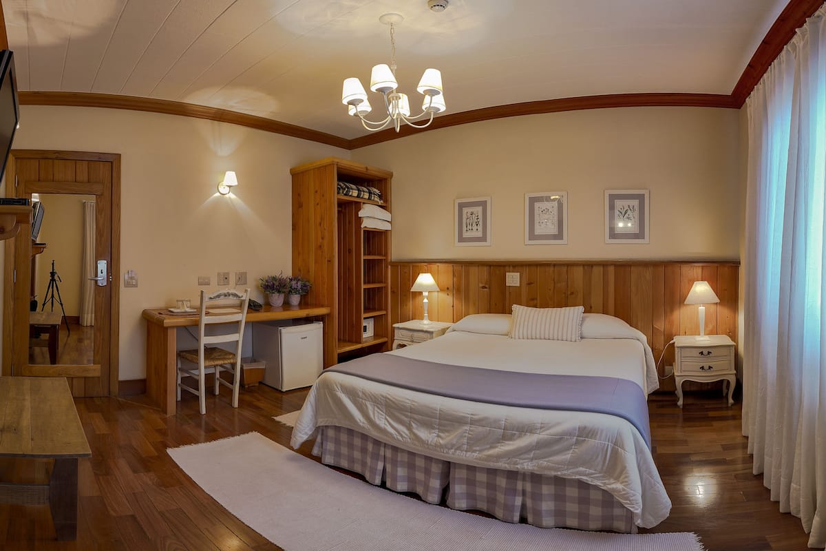 Quarto de hotel com secretária de madeira, cama com colcha xadrez, mesa de cabeceira com abajur e guarda-roupa.