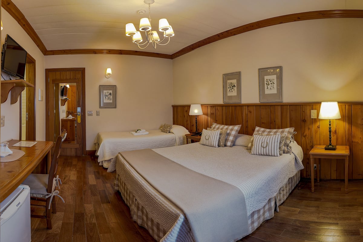 Quarto de hotel com duas camas, piso de madeira, escrivaninha com computador e pequena geladeira.
