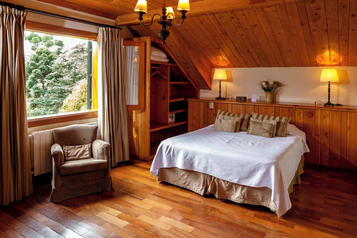 Quarto com teto de madeira, cama com roupa de cama branca, uma cadeira e janela com cortinas.