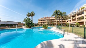Una piscina de borde curvo, rodeada por una valla, palmeras y un edificio con balcones al fondo.