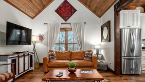 Un salon chaleureux avec un plafond en bois, un canapé en cuir, une table basse en bois agrémentée d’une plante, un téléviseur à écran plat et un réfrigérateur en acier inoxydable.