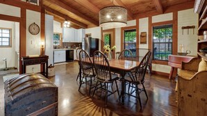 A spacious kitchen with wooden beams, a dining area with a large table and chairs, and a view of the outdoors through large windows. - Cozy Haven | Shared Pool • Firepit • Hot Tub (Fredericksburg)