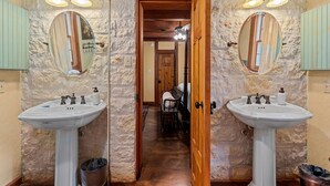 A bathroom with two sinks, each with a mirror and wall-mounted lights. A wooden door leads to a hallway with a ceiling fan and a chair. - Cozy Haven | Shared Pool • Firepit • Hot Tub (Fredericksburg)