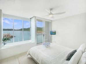 A bedroom with a large window, a ceiling fan, a TV, and a bed with pillows. - 1 Bright Point Townhouse 3103 (Nelly Bay)