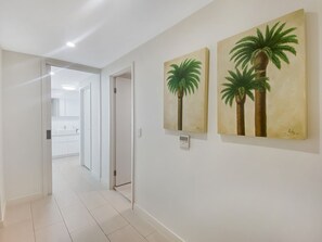 Un couloir aux murs blancs, doté d’un sol carrelé et orné de deux tableaux représentant des palmiers.