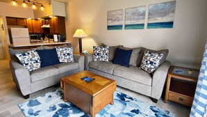 A living room with a grey sofa set, a wooden coffee table, and a blue patterned rug. There is a kitchen area in the background with wooden cabinets and a white refrigerator. - Country Apartment Rental in an Outdoor Paradise! (Mount Carroll)