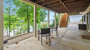 A covered patio with a view of a lake, wooden chairs, and a staircase leading to a building.