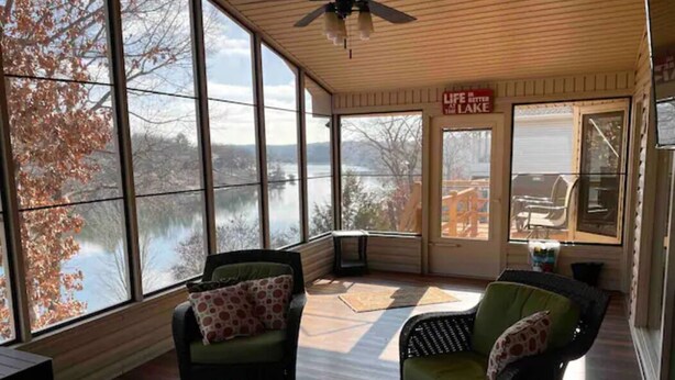 A sunroom with large windows overlooking a lake, furnished with wicker chairs and a glass table.