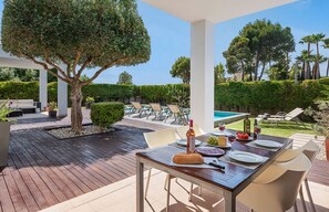 An outdoor dining area with a wooden deck, a table set for two, and a view of a pool and greenery. - Villa Alfa (Puerto Pollensa)