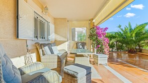 Eine sonnendurchflutete Terrasse mit Korbmöbeln, einem Glastisch und Blick auf einen Garten mit rosa Blumen.