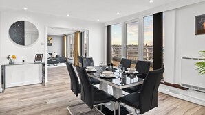 Uma sala de jantar moderna com uma mesa grande posta para seis pessoas, cercada por cadeiras pretas. Um espelho redondo pendurado na parede, e uma janela grande oferece vista para o horizonte da cidade.