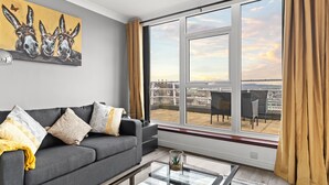 Uma sala de estar moderna com um sofá cinzento, almofadas estampadas, uma mesa de centro de vidro e uma grande janela com vista para o horizonte da cidade.