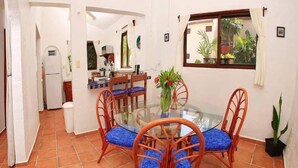 A dining area with a glass table surrounded by wicker chairs with blue cushions, a kitchen in the background, and a window with a view of greenery.
