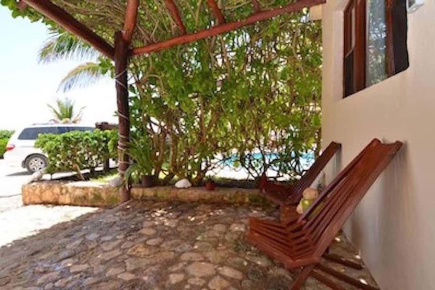 Uma área de estar coberta ao ar livre, com piso de pedra, cadeiras de madeira e um pequeno jardim com árvores e arbustos.