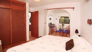 A bedroom with a large bed, a wooden wardrobe, and an arched doorway leading to a dining area with blue chairs and a table.