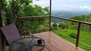 Eine Holzterrasse mit einem Glastisch und einem Stuhl, mit Blick auf eine üppige, grüne Landschaft und ferne Hügel.