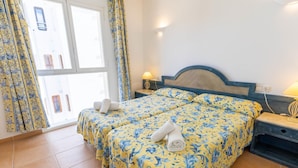 Ein Doppelbett mit floralem Bettbezug, ein Nachttisch mit Lampe und Blick auf ein Gebäude durch ein Fenster.