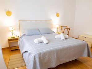 Cama bem arrumada com manta listrada, duas almofadas e dois rolinhos de toalha. Duas mesas de cabeceira com abajures, um criado-mudo de madeira e um pequeno armário de madeira.