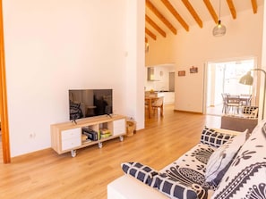 Uma sala de estar com televisão, piso de madeira, sofá e cozinha ao fundo.