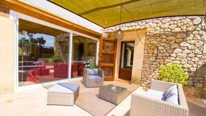 Une terrasse couverte, aménagée avec des meubles en osier, des murs en pierre et une porte vitrée donnant sur un espace intérieur.