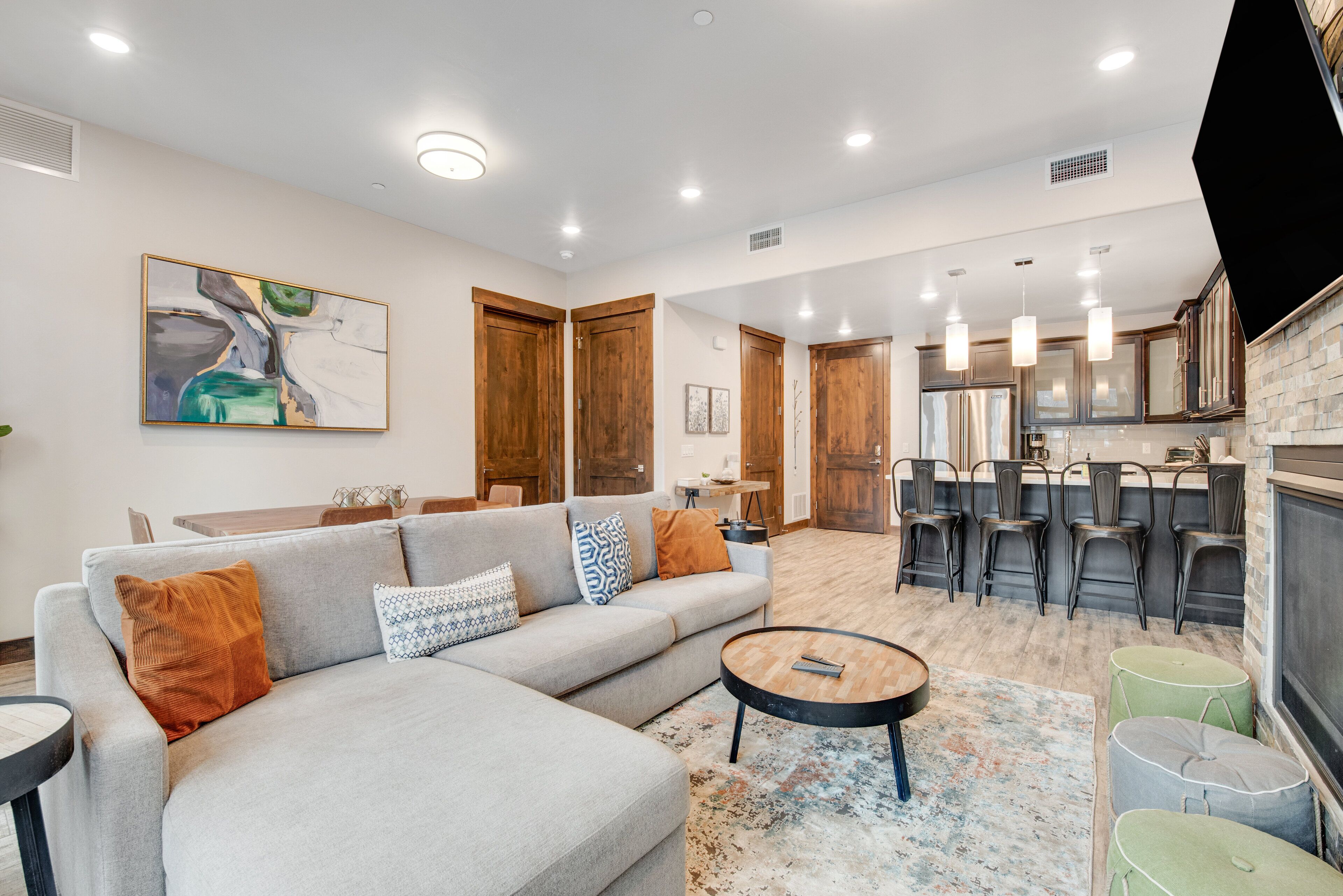A modern living room with a sofa, coffee table, and ottomans. There is a kitchen area with bar stools and a dining table.