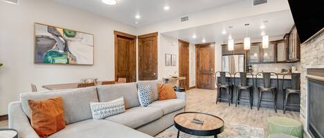 A modern living room with a sofa, coffee table, and ottomans. There is a kitchen area with bar stools and a dining table.