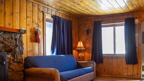 Ein gemütliches Blockhauskaminzimmer mit Holzbalkendecke, Steinwand, blauem Sofa, einer Lampe und Kamin.