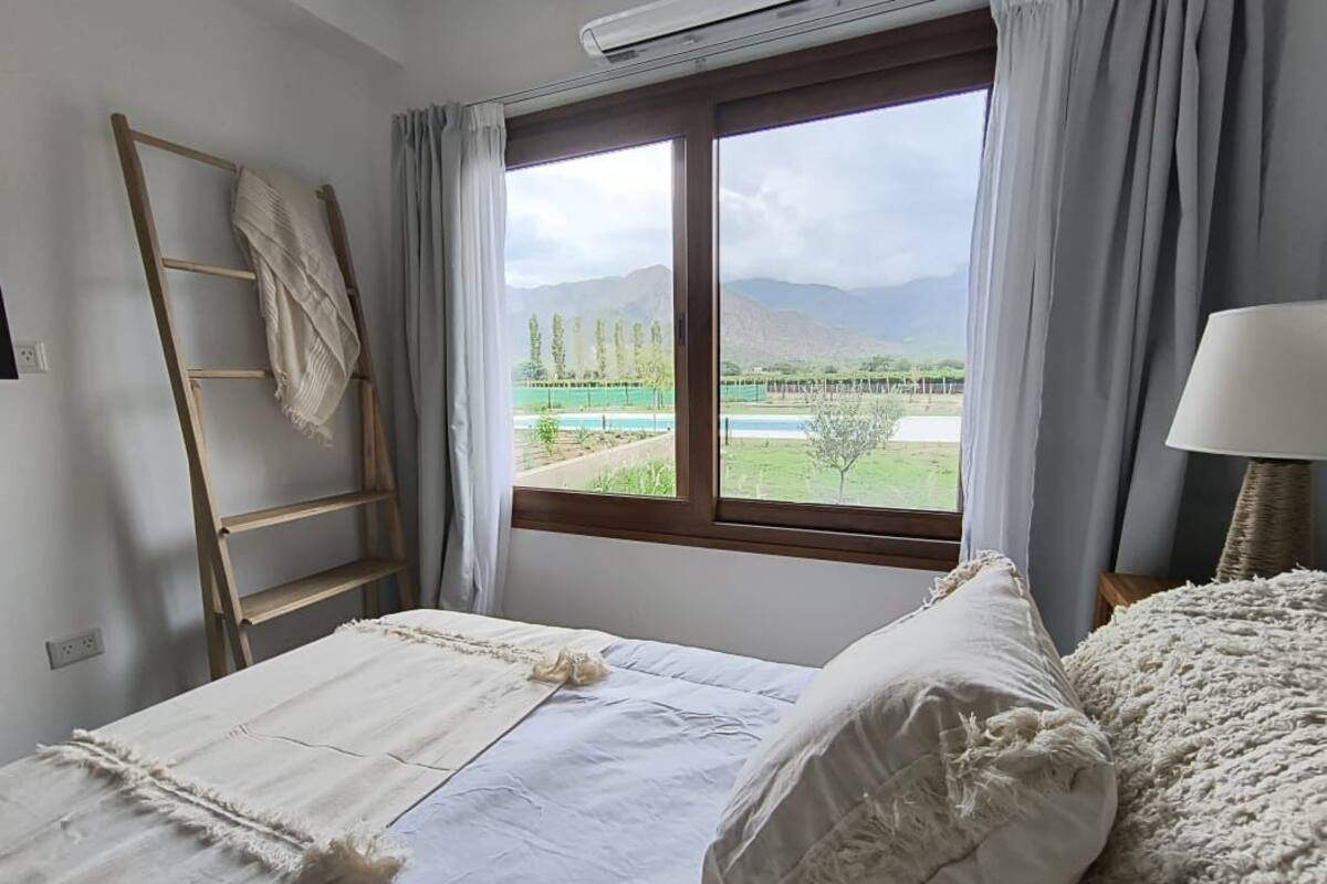 Quarto com cama, escada, janela com cortinas e vista para a paisagem.