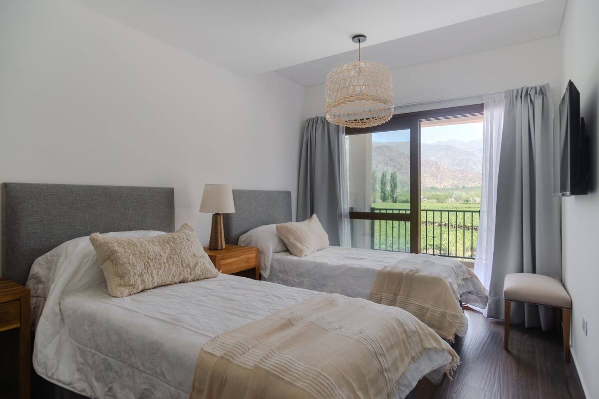 Quarto com duas camas, mesa de cabeceira com abajur, TV na parede e varanda com vista para as montanhas.