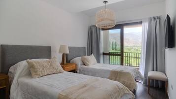 Un dormitorio con dos camas, una mesita de noche con lámpara, un televisor montado en la pared y un balcón con vistas a las montañas.