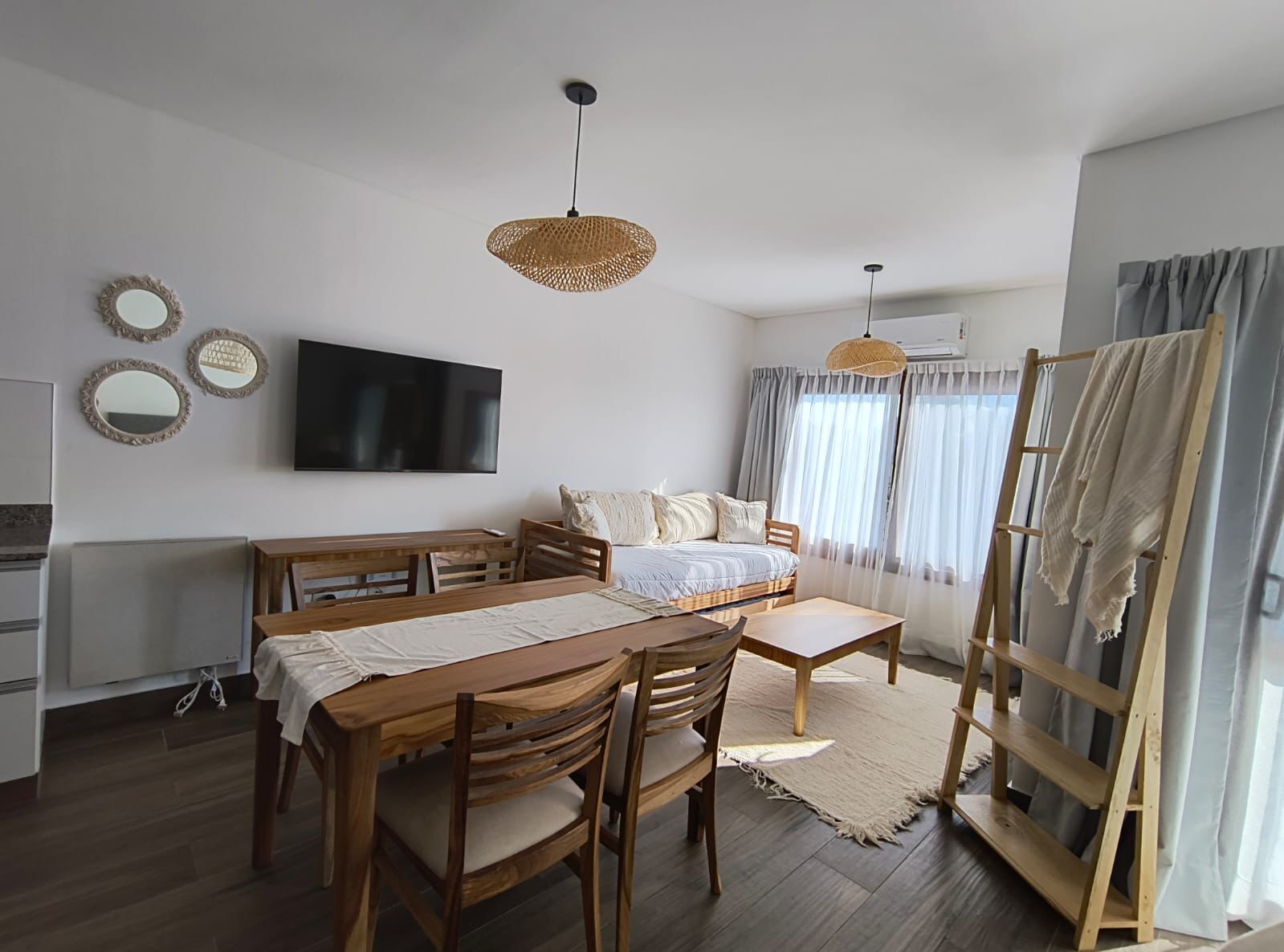 Una habitación moderna y bien iluminada con una mesa de comedor de madera, sillas, un sofá y un televisor montado en la pared.