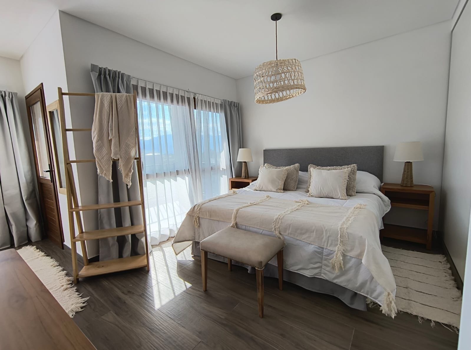 Un dormitorio con cama, mesitas de noche, una lámpara colgante, una escalera de madera y vistas al exterior a través de cortinas translúcidas.