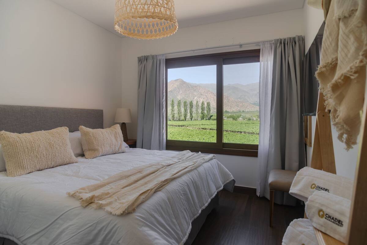Quarto com uma cama grande, cabeceira de madeira, janela com vista para um campo verde e montanhas, e luminária suspensa.
