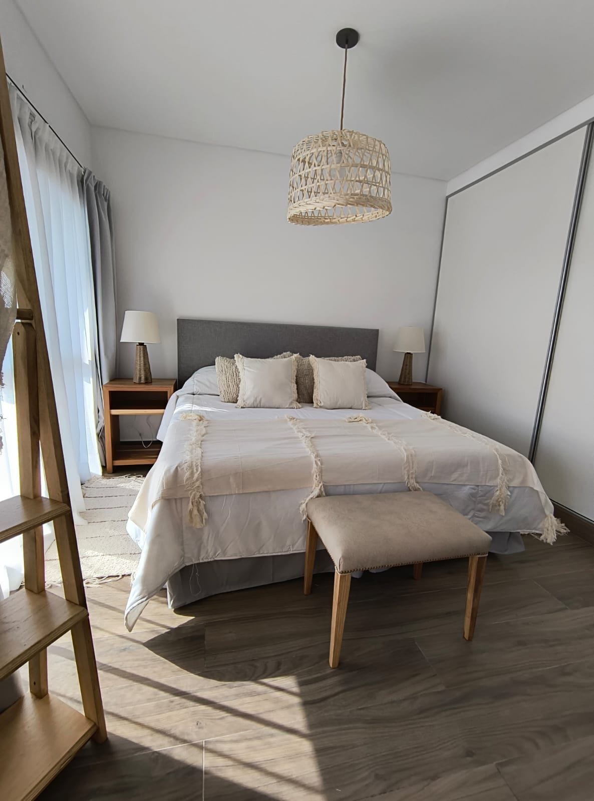 A bedroom with a large bed, a wooden bench, a nightstand, a lamp, and a woven ceiling light fixture.