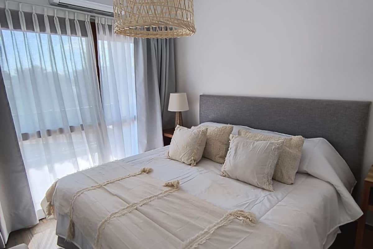 Quarto moderno com cama grande, luminária pendente de palha e vista para o mar através de cortinas translúcidas.
