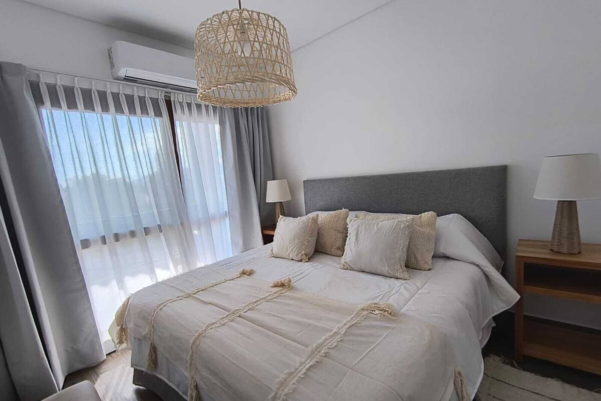 Quarto com uma cama grande, uma mesa de cabeceira com abajur, um candeeiro tecido pendente e uma janela com cortinas.