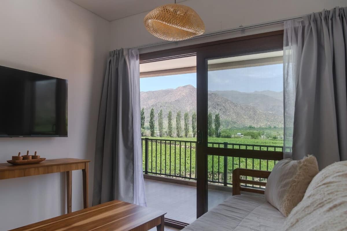 Quarto com cama, televisão presa na parede, mesa de madeira e varanda com vista para montanhas.