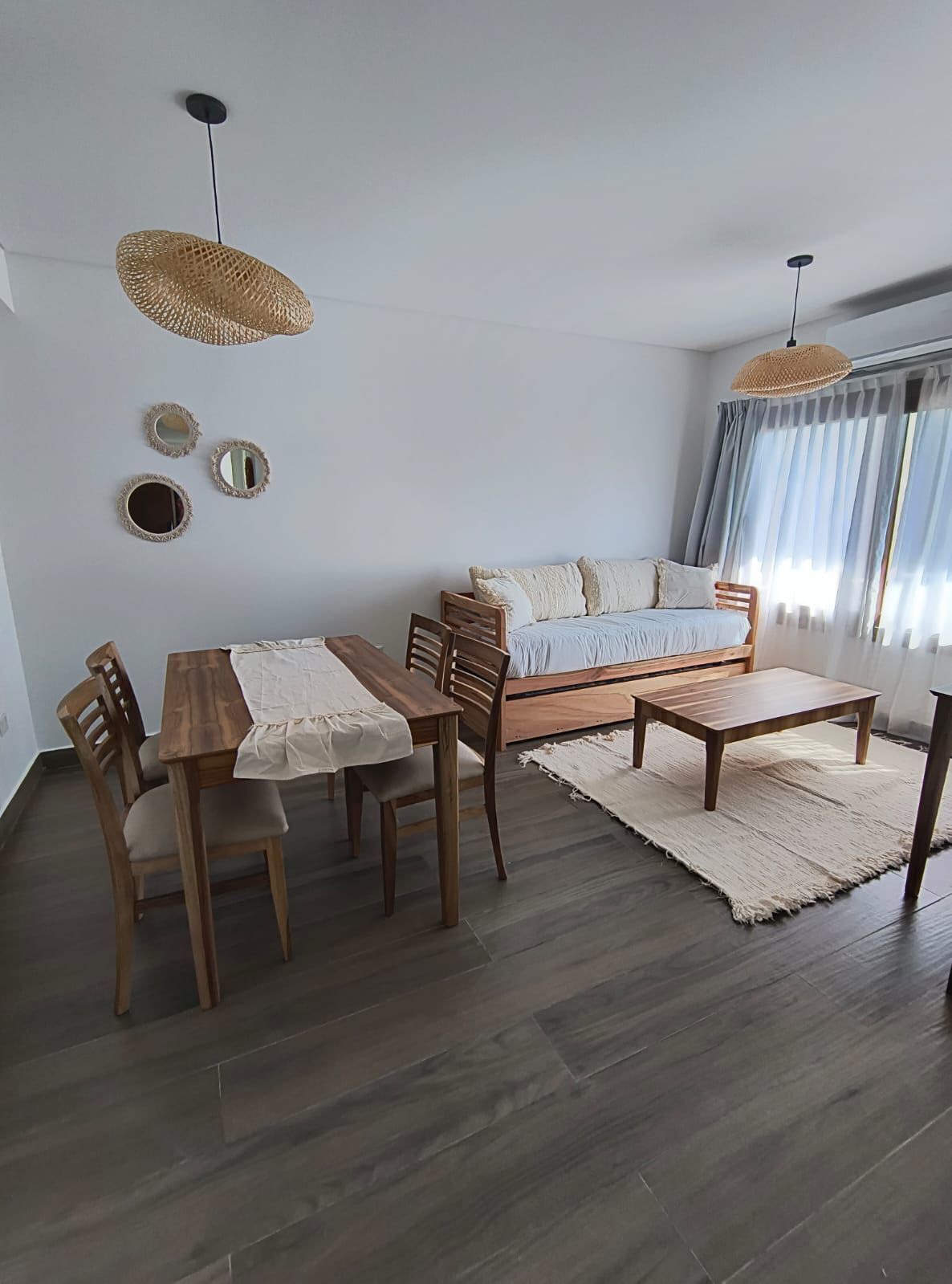 A modern living room with a wooden dining table, chairs, a sofa, and a large window with curtains.
