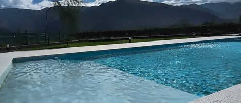 A swimming pool with clear blue water, surrounded by a white deck. In the background, there are mountains and a partly cloudy sky.