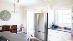 A modern kitchen with a stainless steel refrigerator, white cabinetry, a dining area with wooden chairs, and a decorative wall piece.