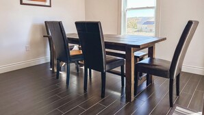 A dining area with a wooden table and four chairs, a pendant light, a window with a view, and a framed picture on the wall.