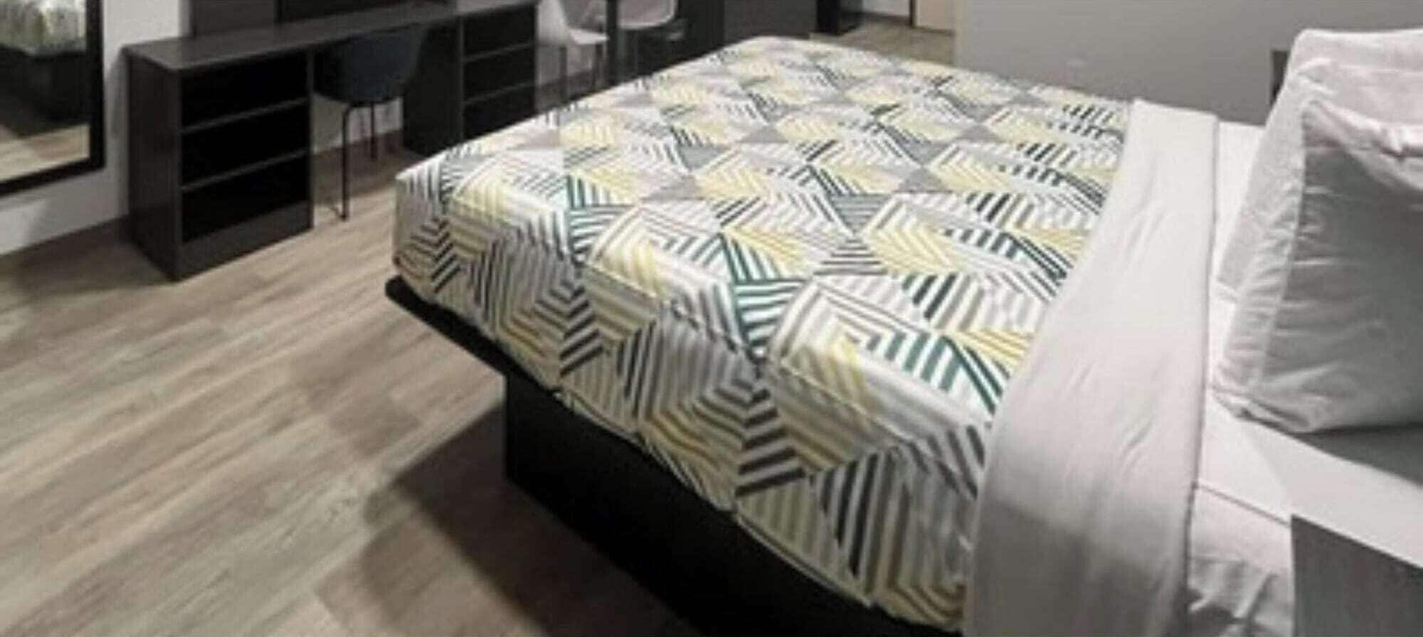 Een net opgemaakt bed met een deken met een geometrisch patroon, een donker hoofdbord en een wit kussen.