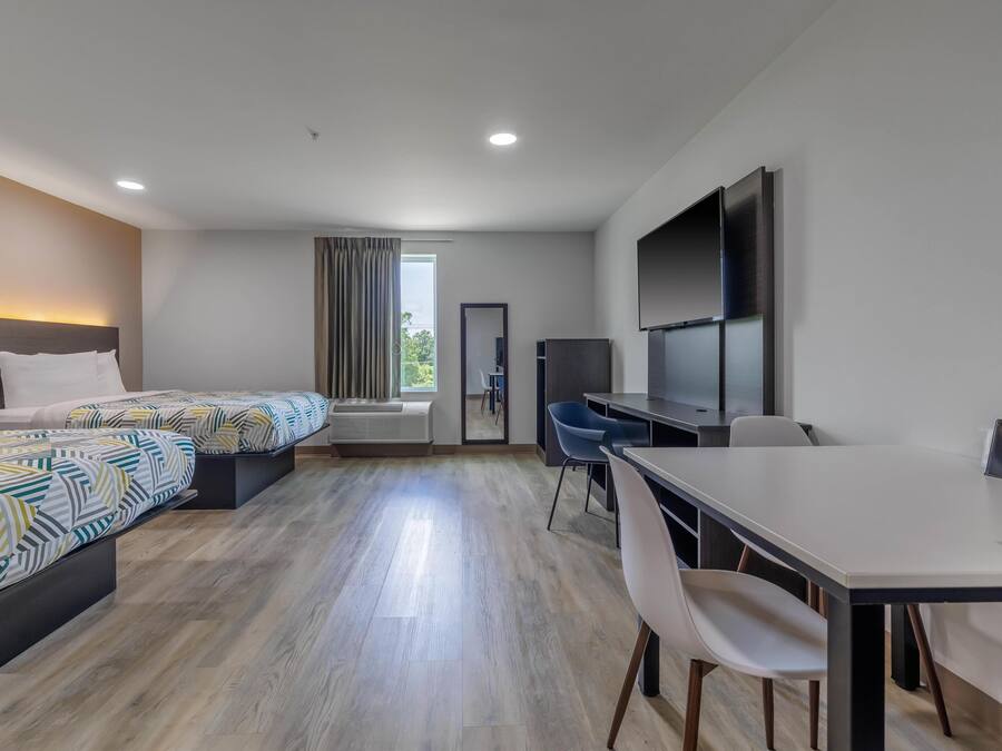 A hotel room with two beds, a desk, chairs, a TV, and a large window.