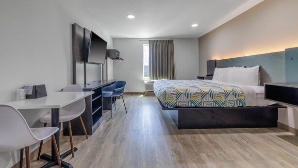 A modern hotel room with a bed, desk, chairs, and a TV. - Studio 6 Gonzales, La - Baton Rouge/Geismar/Prairieville I-10 (Gonzales)